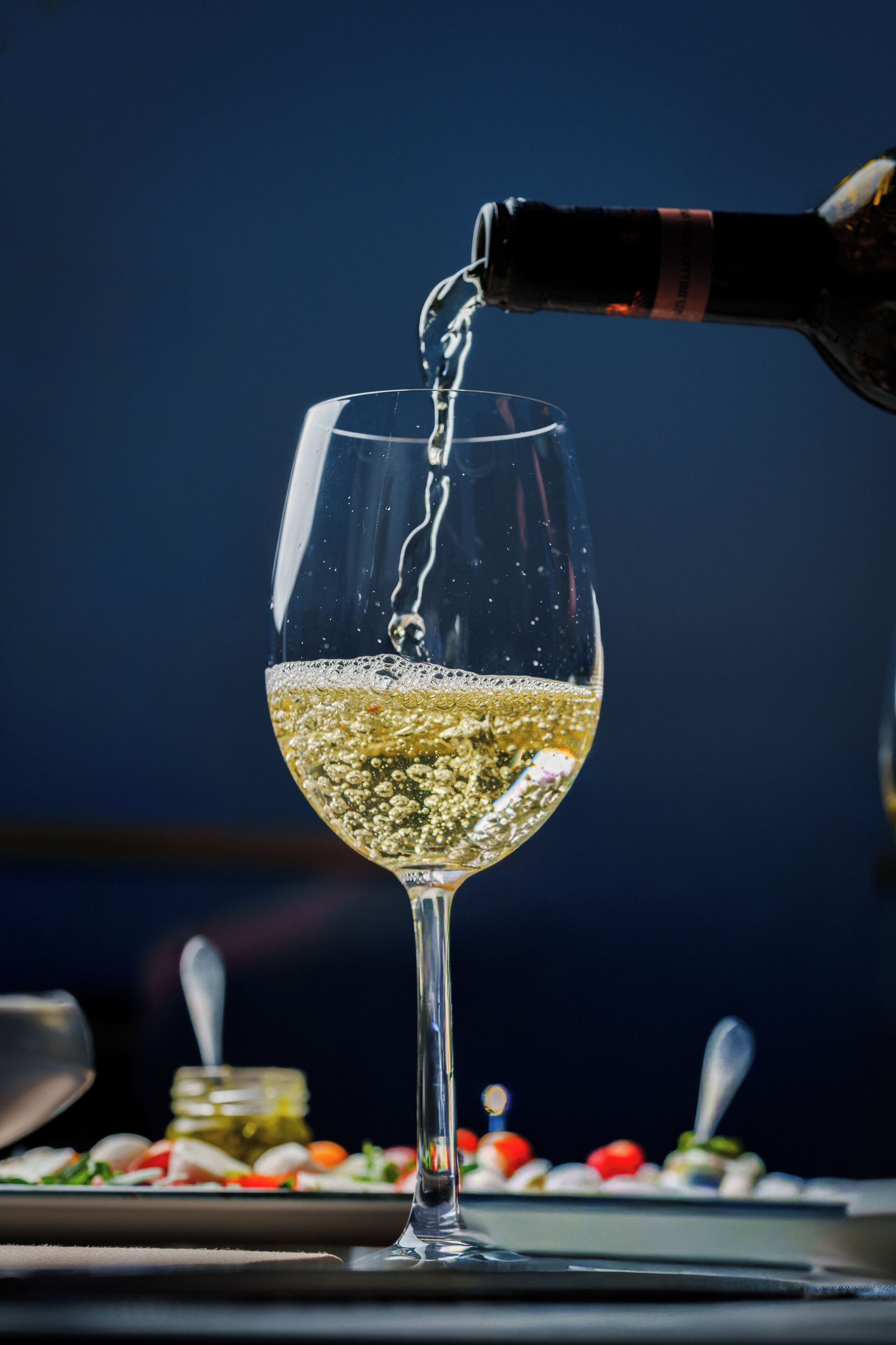 A glass of white wine being poured, with a dark background and a plate of food in the foreground.