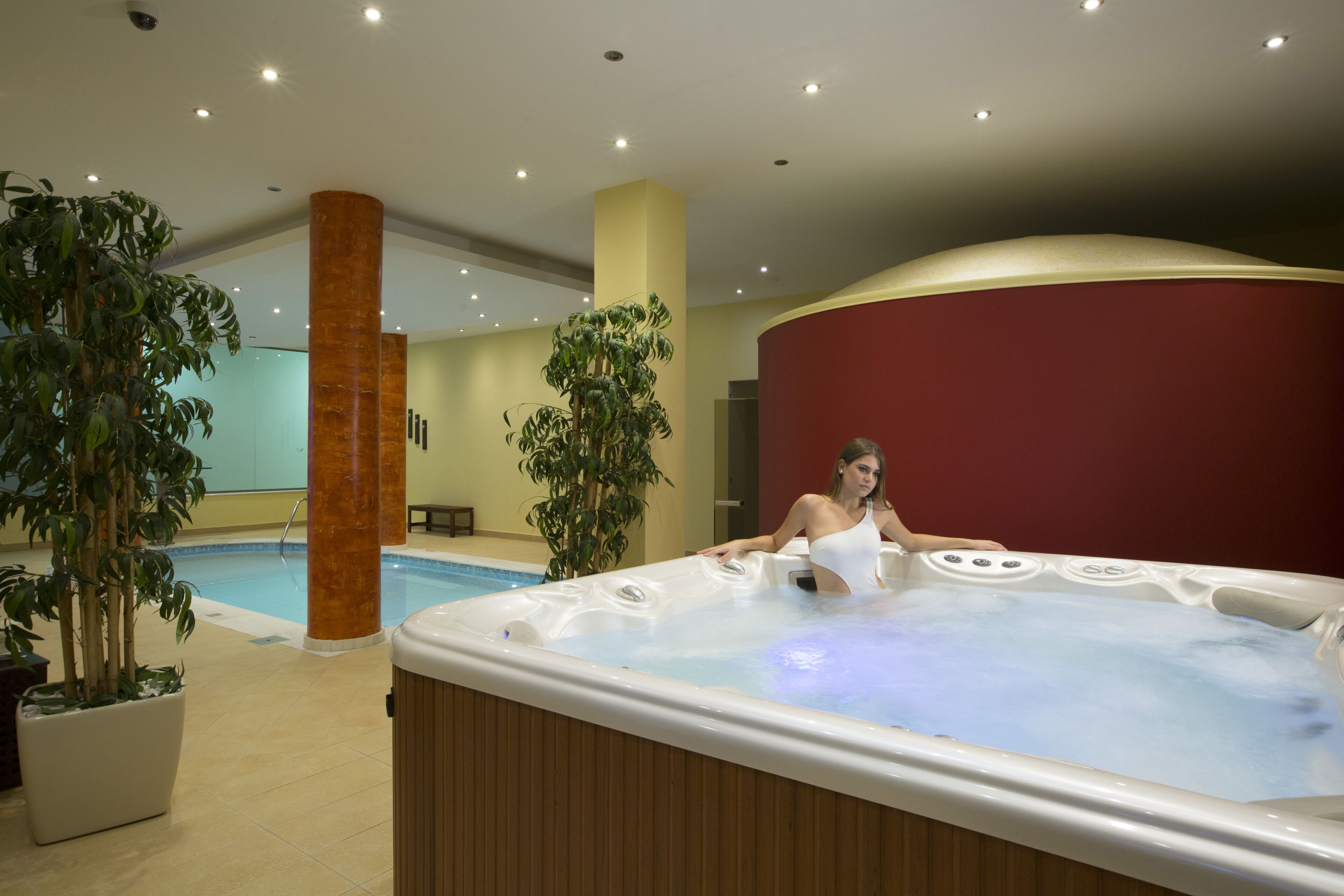 A hot tub with a person relaxing in it, surrounded by indoor plants and a pool.