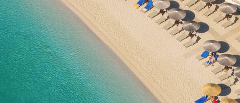 An aerial view of a beach with rows of sun loungers and umbrellas, a clear blue sea, and a sandy shore.