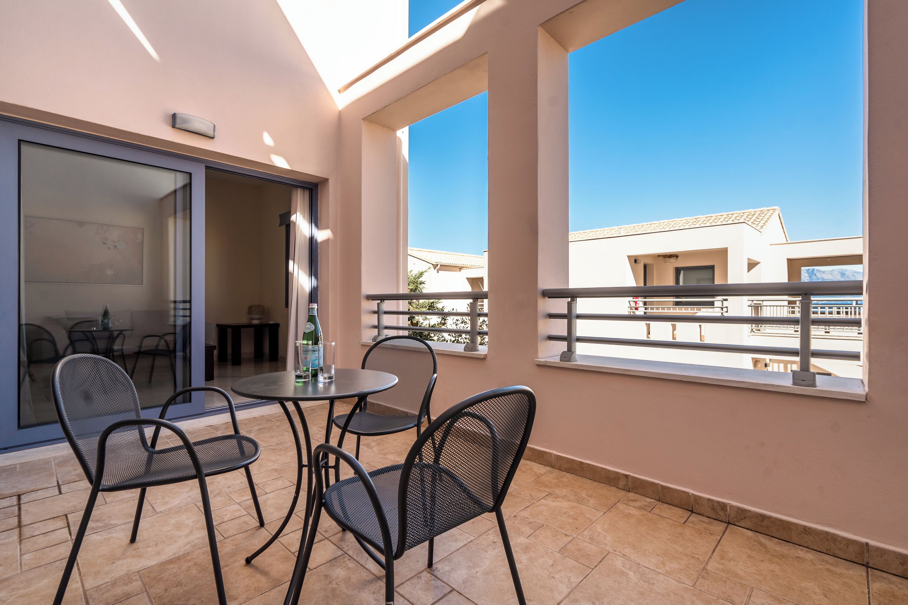 Ein Balkon mit Tisch und Stühlen, eine Glastür, die zu einem Innenbereich führt, und Blick auf Gebäude im Hintergrund.