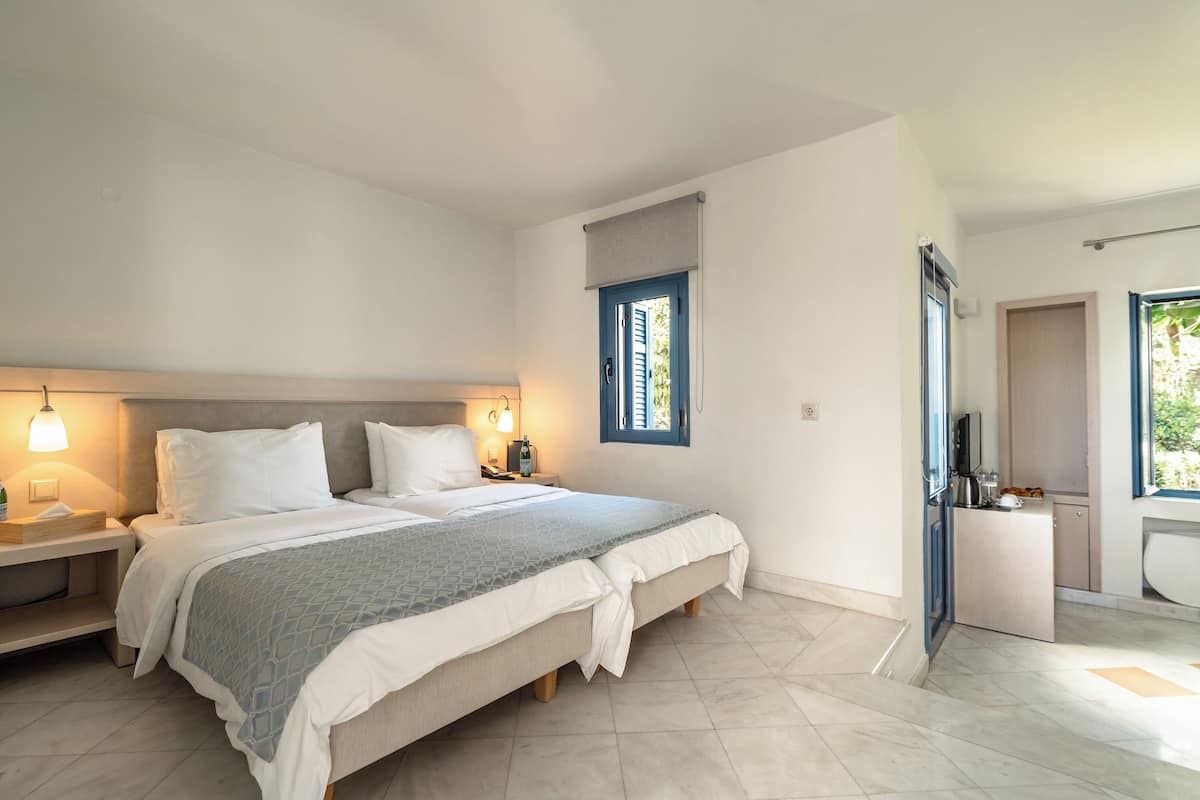 A bedroom with a bed, bedside tables, a nightstand, a window with shutters, and a door leading to a bathroom.