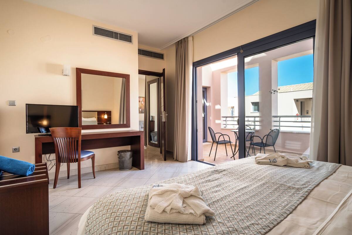 A hotel room with a bed, a desk with a chair, a television, and a balcony with chairs.