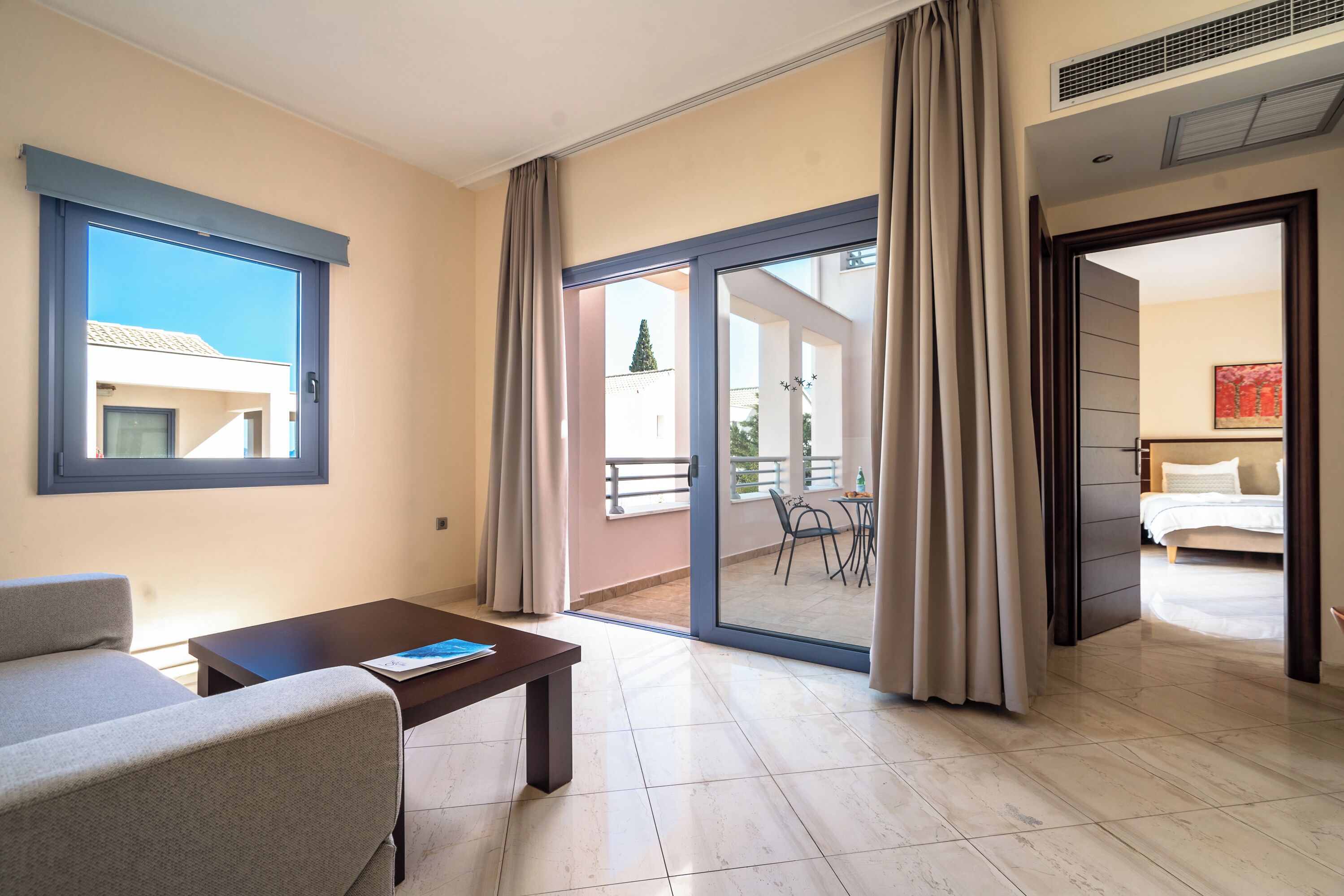 A modern hotel room with a sofa, a small table, a balcony with a view of the outside, and a sliding door leading to another room.