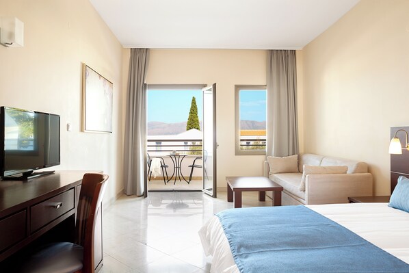 A hotel room with a bed, a desk with a TV, a balcony with a table and chairs, and a view of the mountains. - Porto Galini Seaside Resort & Spa (Lefkada)