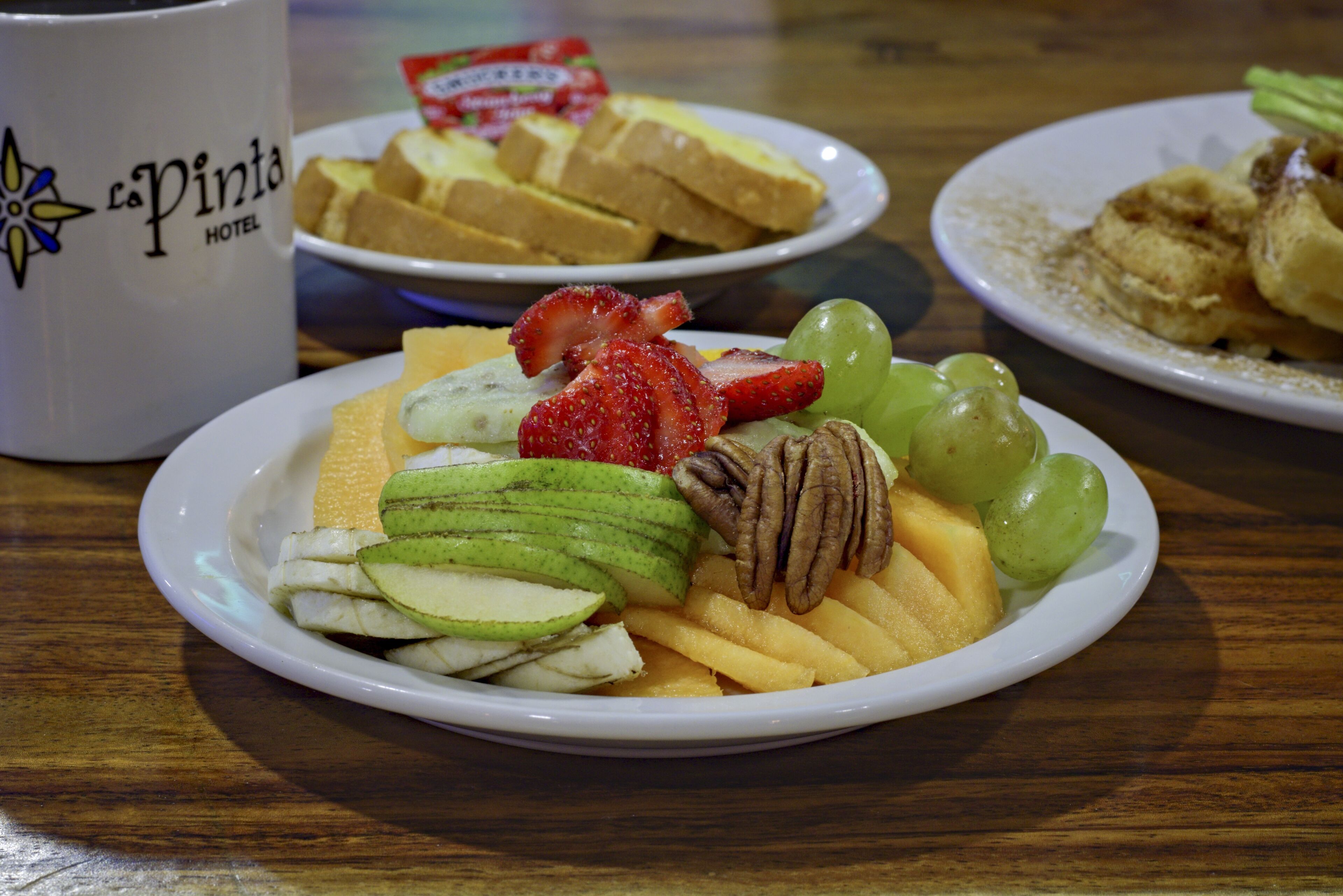 A plate of fruit including strawberries, kiwi, and grapes, with a white mug labeled "La Pinta Hotel" in the background.