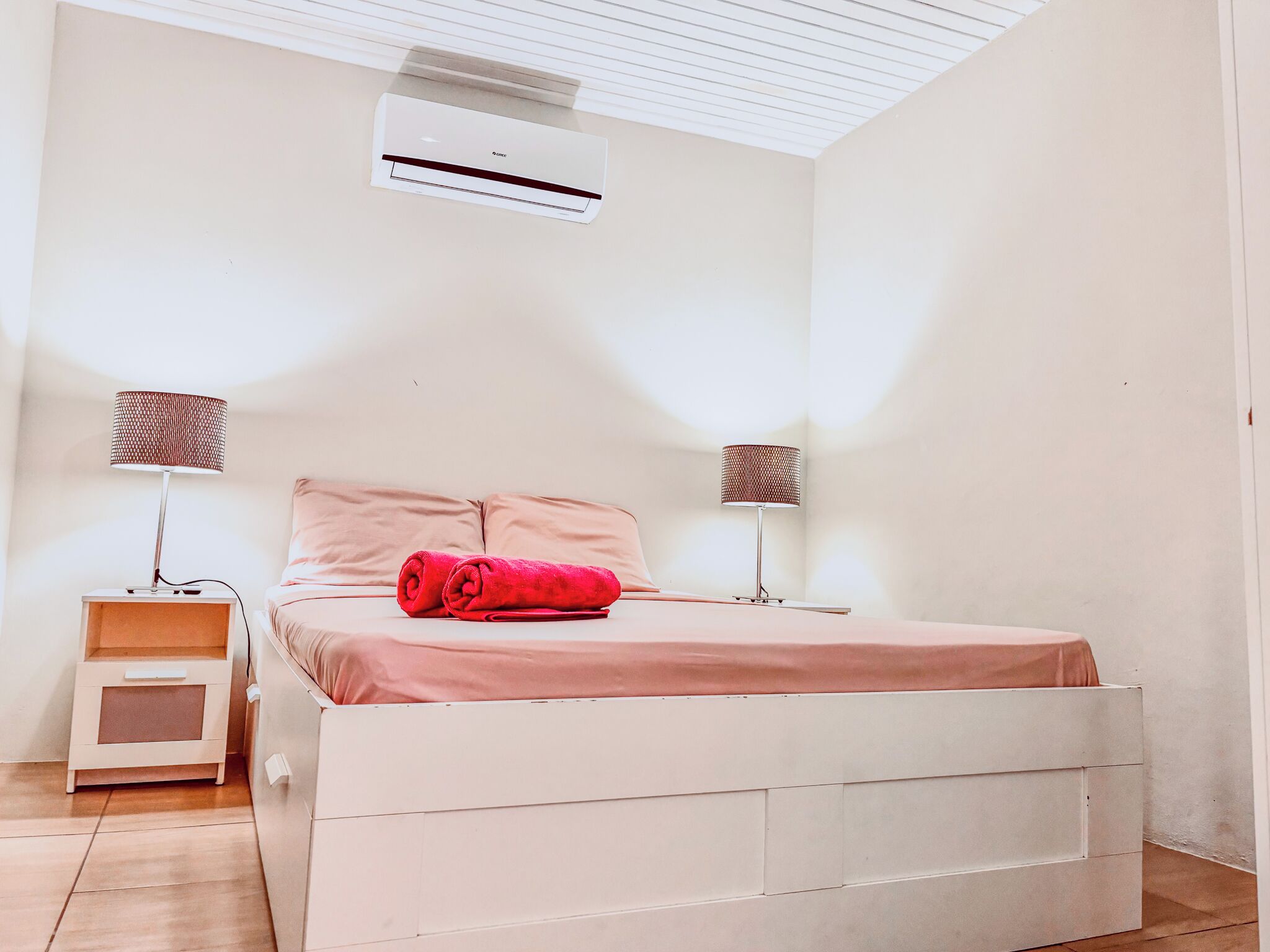 A bedroom with a bed, two bedside tables, an air conditioner, and a lamp on each side.