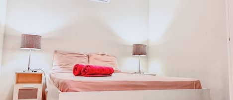 A bedroom with a bed, two bedside tables, an air conditioner, and a lamp on each side.