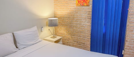 A hotel room with a bed, a nightstand with a lamp, a brick wall, and a blue curtain.
