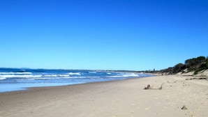 Una playa con cielo despejado, orilla de arena y vegetación escasa a la derecha.