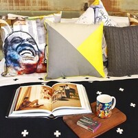 Una cama con un libro y una taza en una bandeja, almohadas con un diseño geométrico y una manta con un motivo de cruz.
