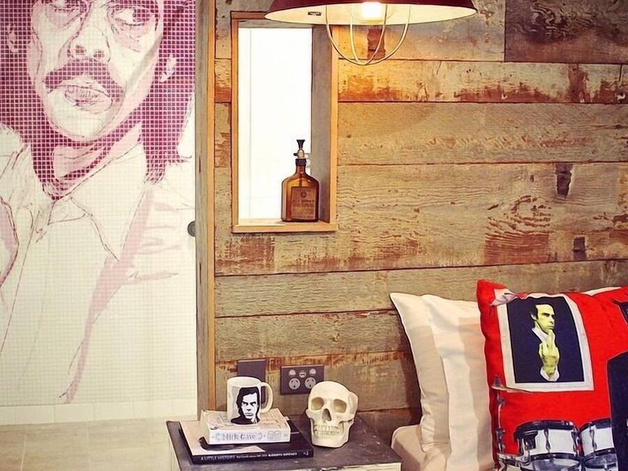 A bedroom with a wooden wall, a bed with red and black pillows, a nightstand with a skull and books, and a poster of a man.