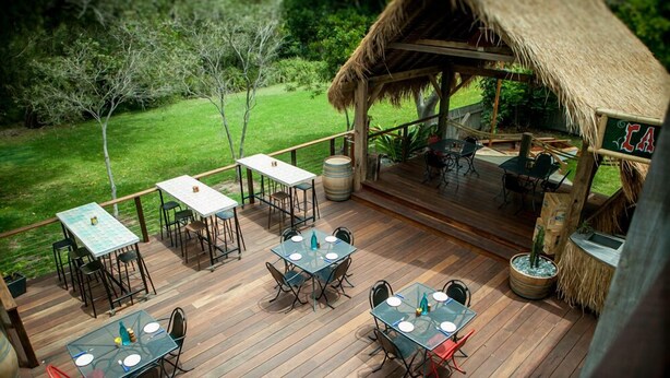 Una terraza al aire libre con piso de madera, varias mesas y sillas, una estructura de techo de paja y vistas a un paisaje verde.