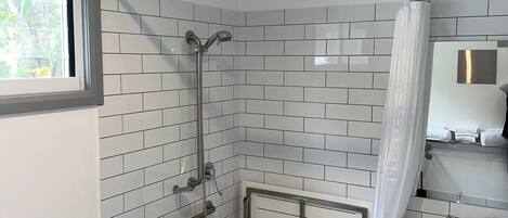 A modern bathroom with white subway tiles, a shower with a glass door, a handrail, a sink with a mirror, and a window with a view of greenery.
