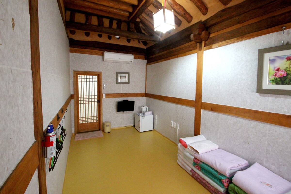 Ein Zimmer mit Holzbalken, einem gelben Boden, einem Bett mit Kissen, einem kleinen weißen Heizgerät und einem Bild an der Wand.