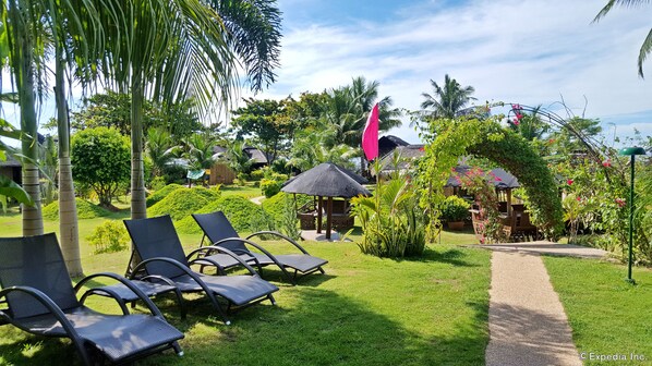 Un resort tropical con estructuras de techo de paja, palmeras y un césped cuidado con sillas de descanso.