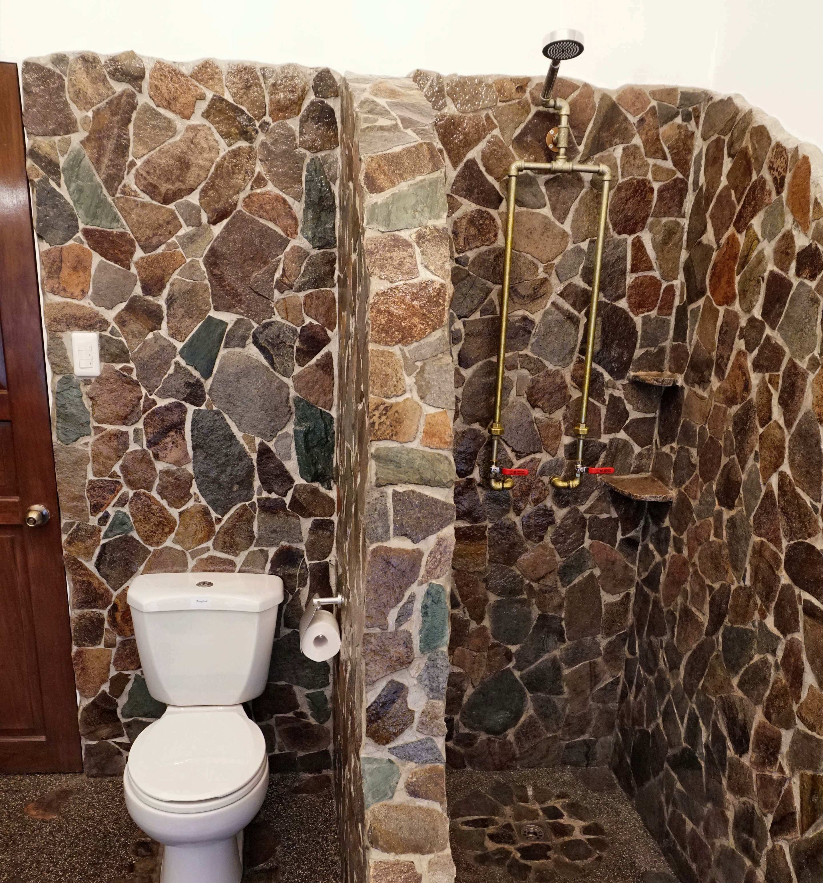Ein Badezimmer mit einer Steinwand, einer Toilette und einer Dusche mit einem Metall-Duschkopf.