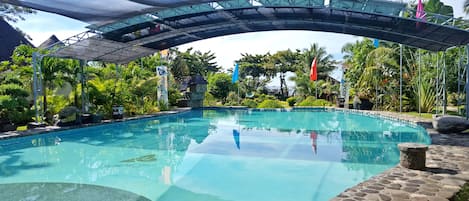 Piscina cristalina con borde de piedra, rodeada de exuberante vegetación y un toldo sombreado.