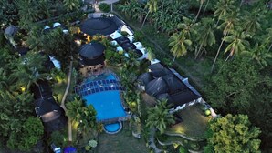 An aerial view of a resort with a central swimming pool, multiple covered areas, and lush tropical vegetation.
