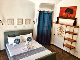 A neatly made bed with a decorative swan towel, a wall-mounted air conditioner, a blue curtain, a wall art of a tree, and a wooden hanger rack.