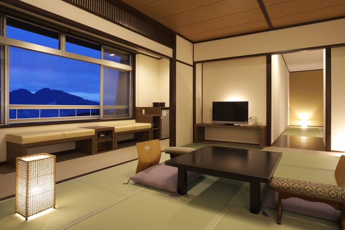 A traditional Japanese room with tatami flooring, a low table, seating, a television, and a view of mountains.