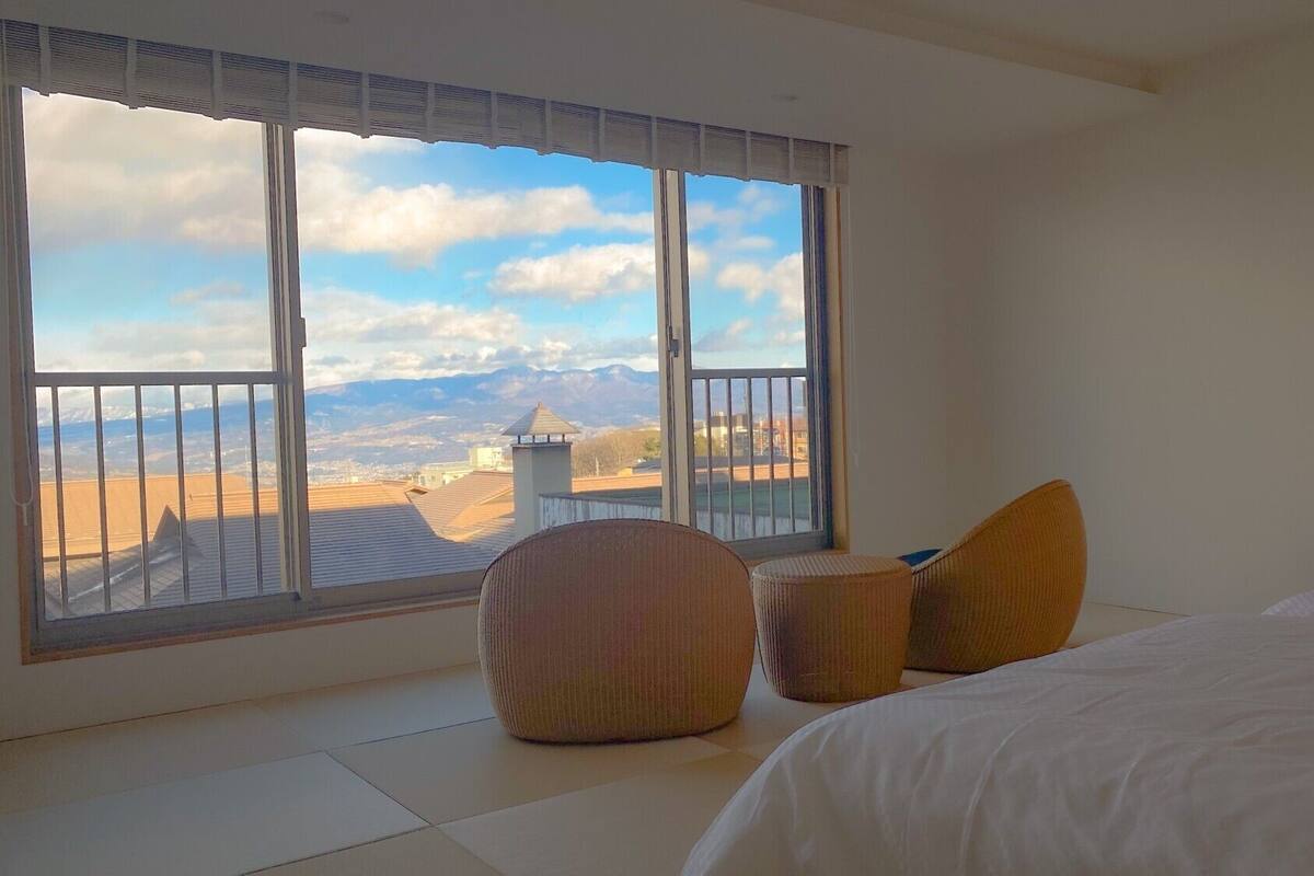 A bedroom with a large window, a bed, and two ottomans.