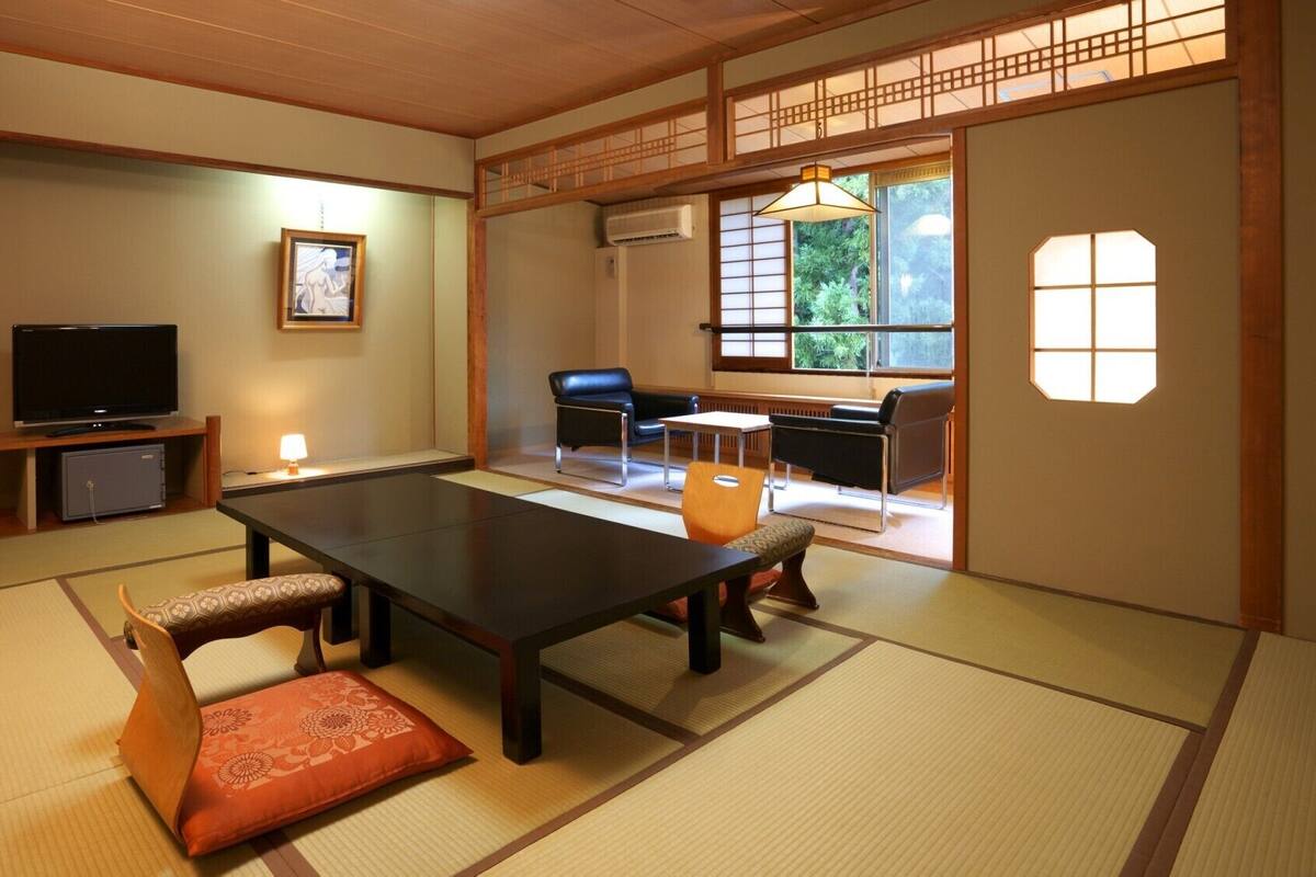 A traditional Japanese room with tatami flooring, a low table with cushions, a television, and a sliding door leading to a seating area with a view of greenery.
