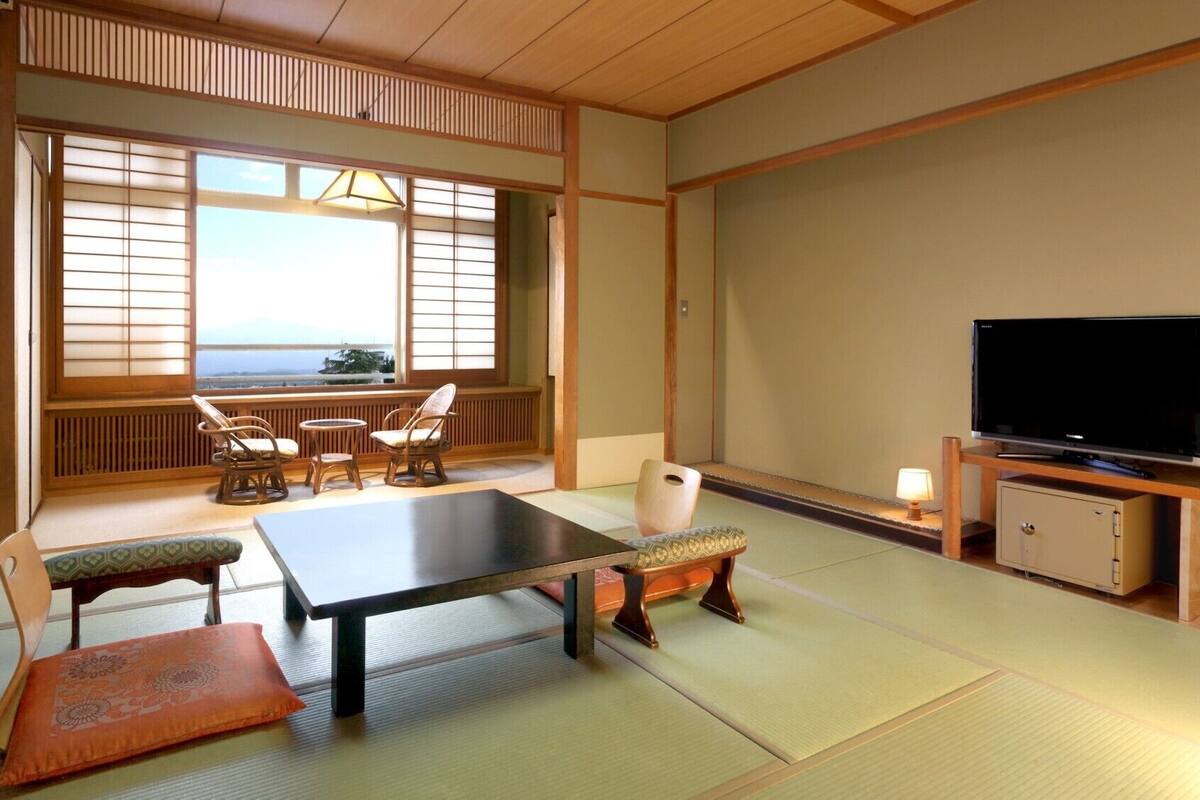A traditional Japanese-style room with tatami flooring, a low table, chairs, a television, and a view of the ocean.