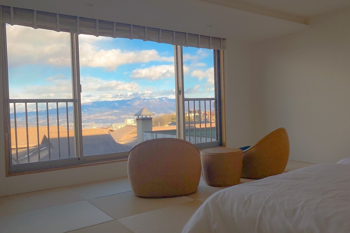 A bedroom with a large window, a bed, and two ottomans.