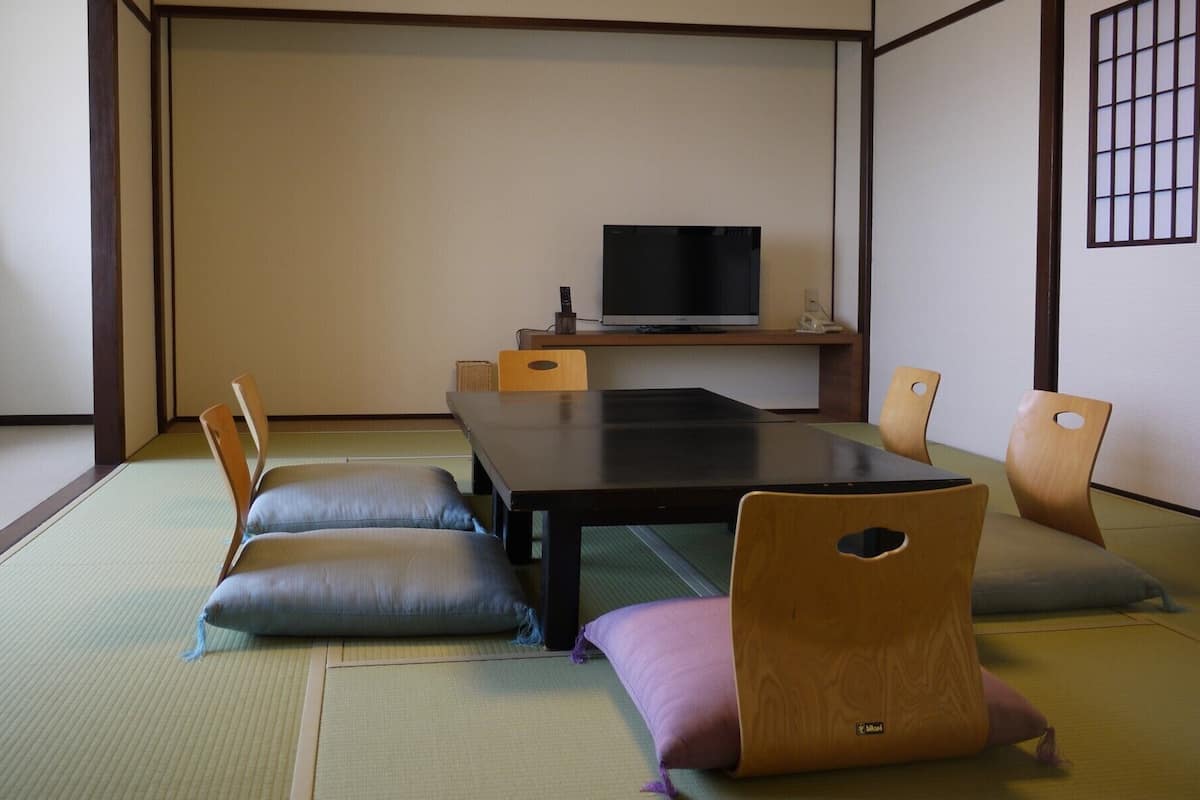 Ein Raum mit Tatami-Matten, einem niedrigen Tisch, Kissen, einem Fernseher und einem kleinen Regal.