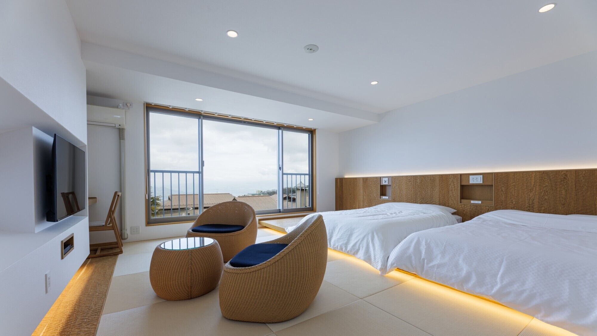 A modern hotel room with a large balcony, two beds, a TV, and wicker chairs.