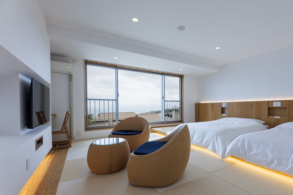 A modern hotel room with a large balcony, two beds, a TV, and wicker chairs.