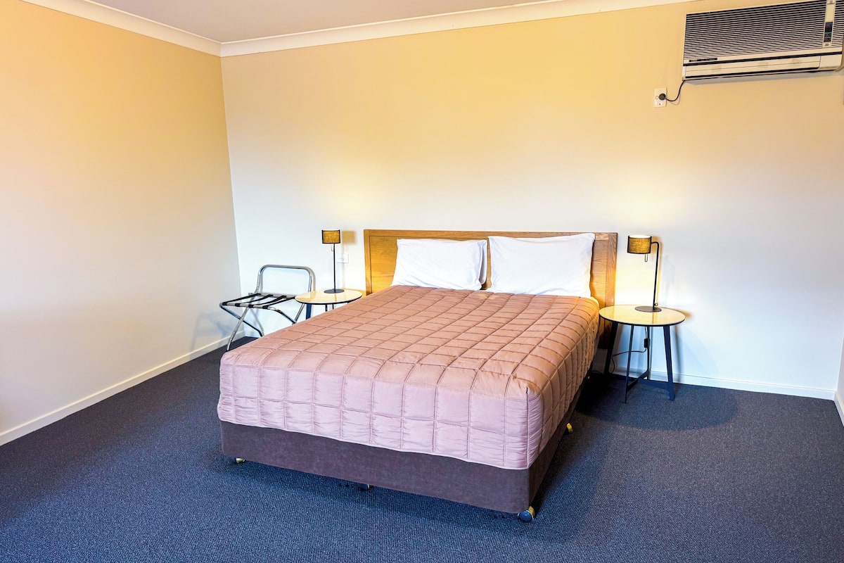 A hotel room with a large bed, two bedside tables, a chair, and a wall-mounted air conditioning unit.
