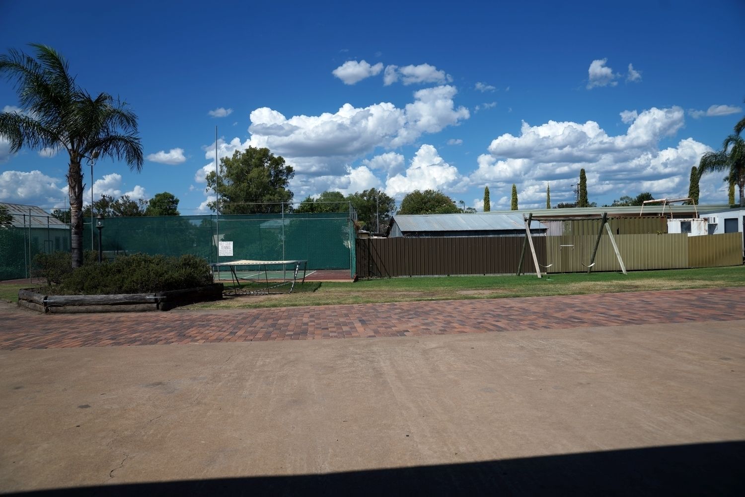 A tennis court with a green fence, a palm tree, and a building with a metal roof.