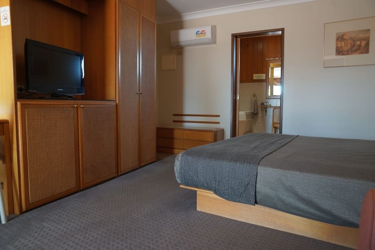 A bedroom with a bed, a television on a wooden cabinet, a wardrobe, and a bathroom visible through an open door.
