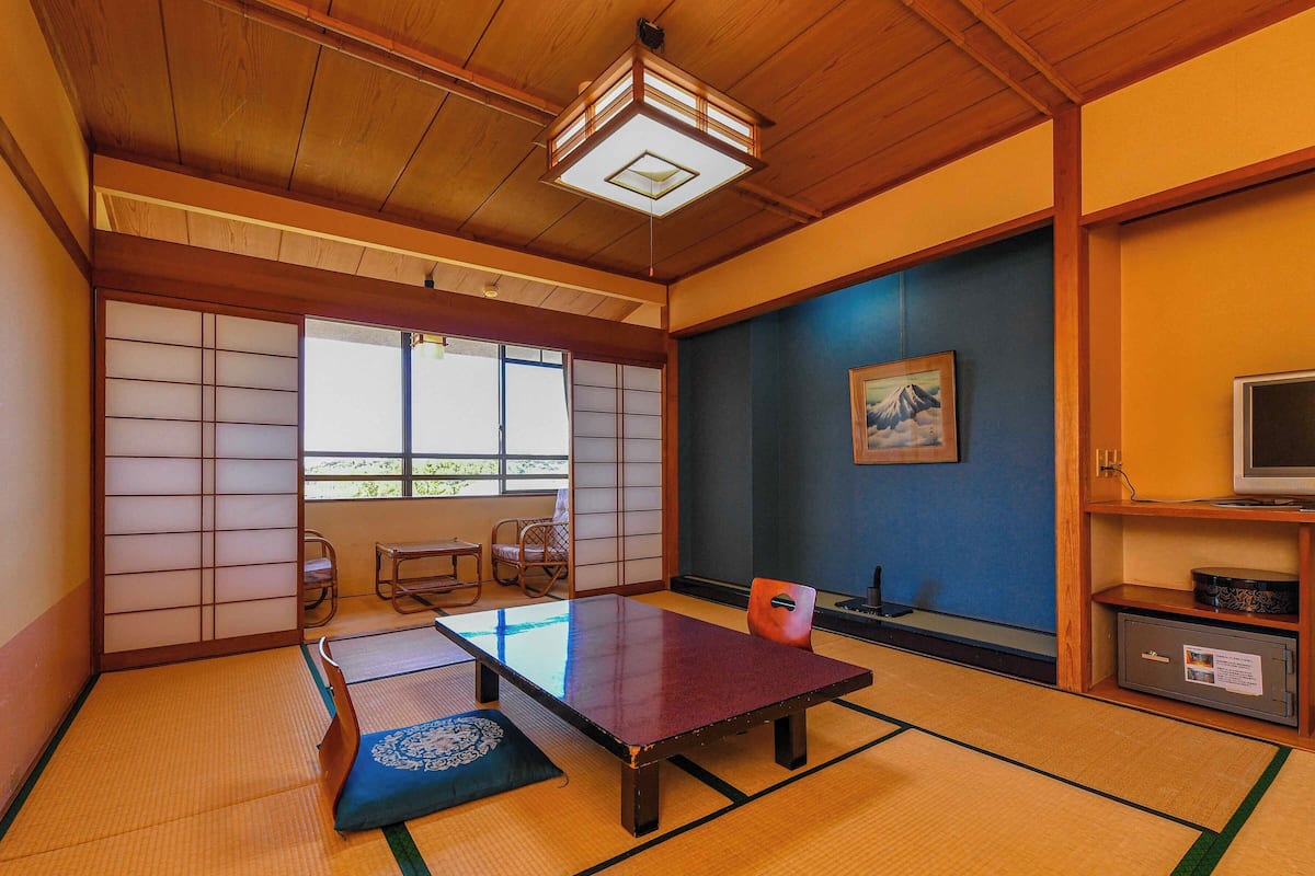 A traditional Japanese room with tatami flooring, a low table, a cushioned bench, and a television on a wooden shelf.