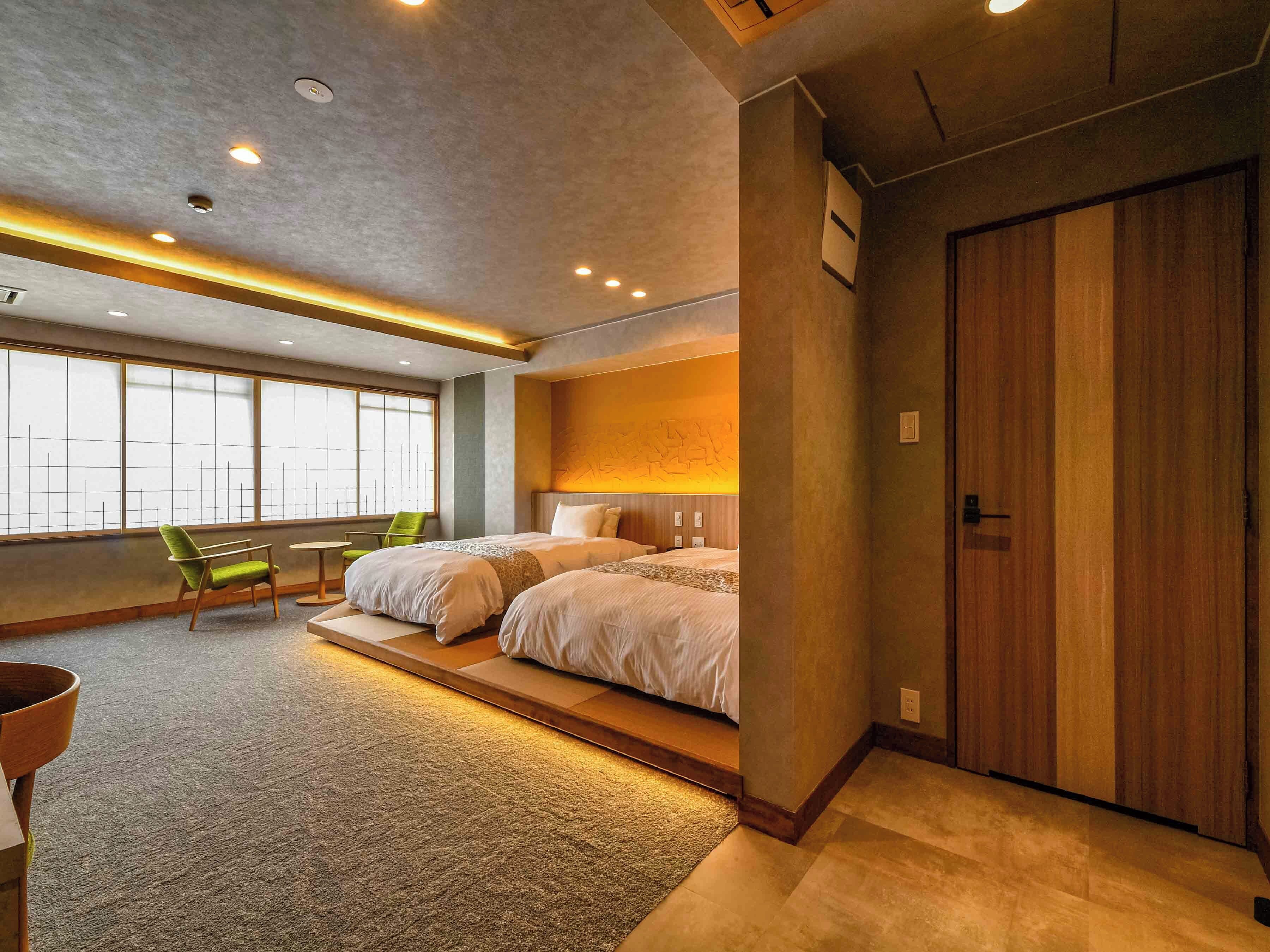 Una habitación de hotel moderna con dos camas, una silla verde, una mesita pequeña y un ventanal grande.