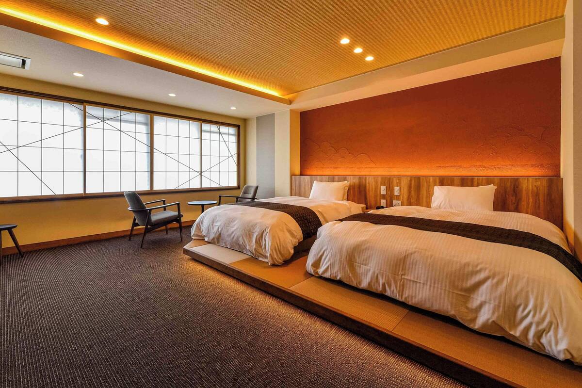 A modern hotel room with a large bed, a seating area with chairs, and a large window providing a view.