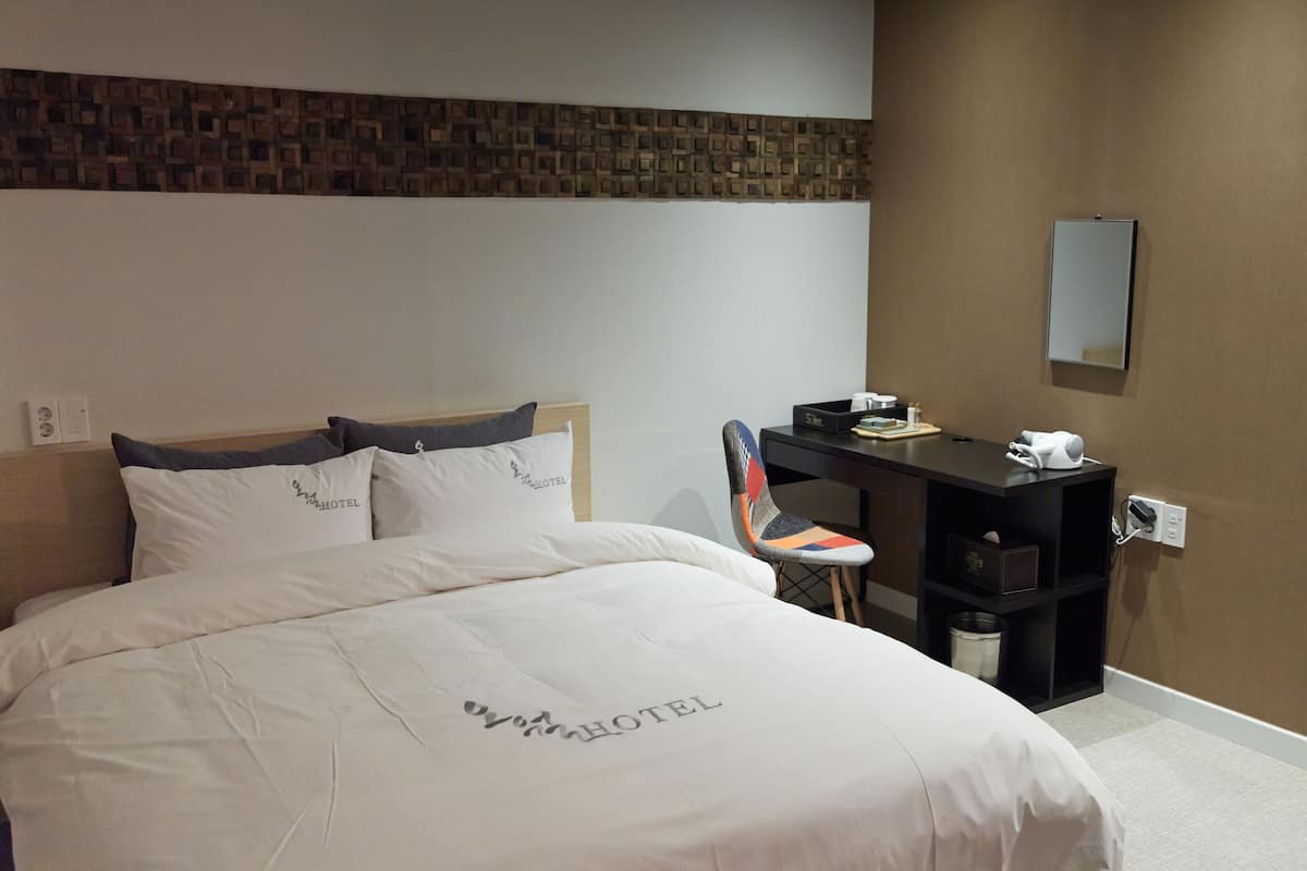 A hotel room with a neatly made bed, a desk with a chair, a mirror, and a wall-mounted shelf.