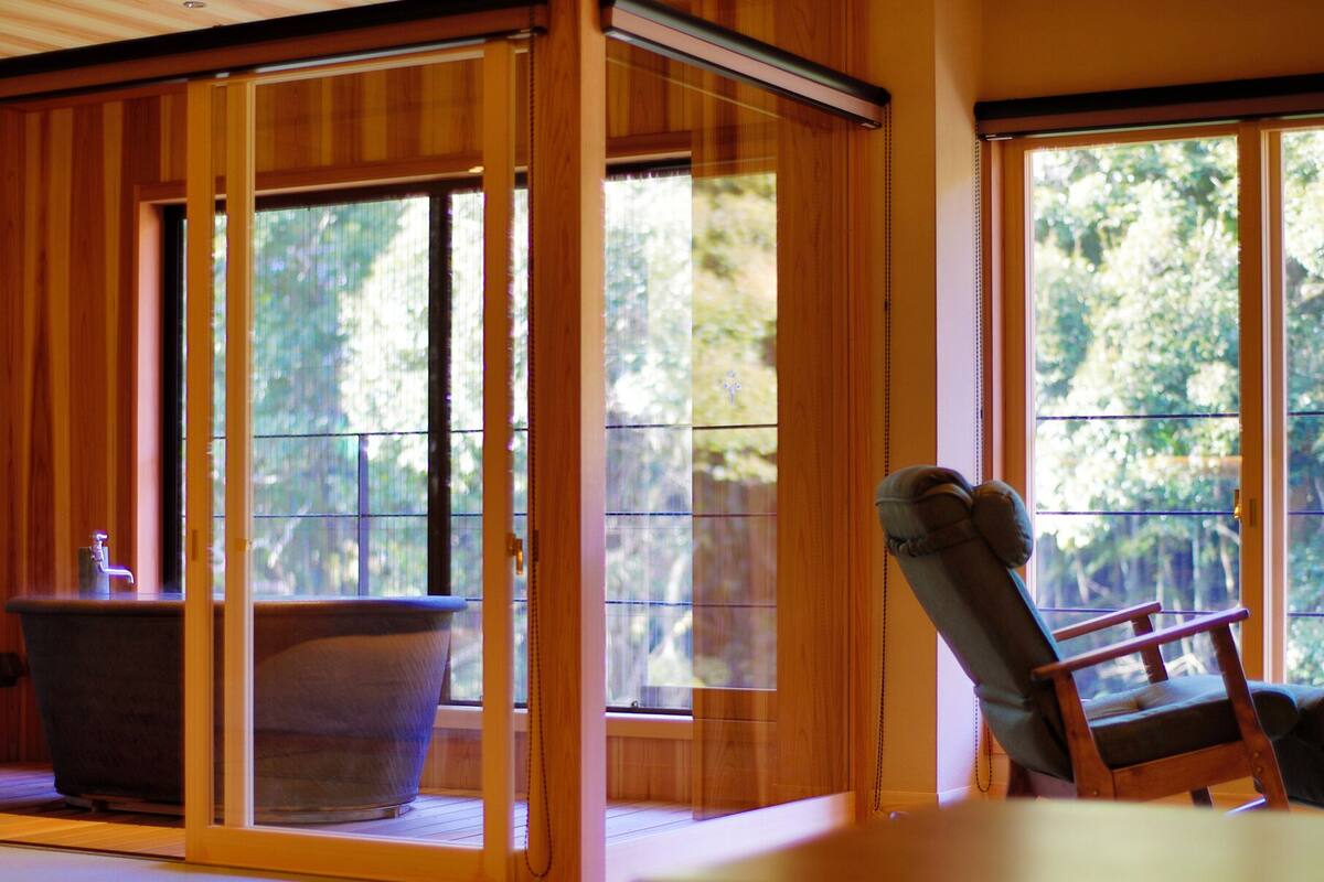 Una habitación con puertas correderas de madera, una bañera y una silla junto a una ventana con vistas a árboles.