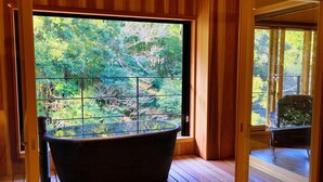 A wooden interior with a large window, a bathtub, and a view of greenery outside. - Tatsuta Ryokan (Izu)