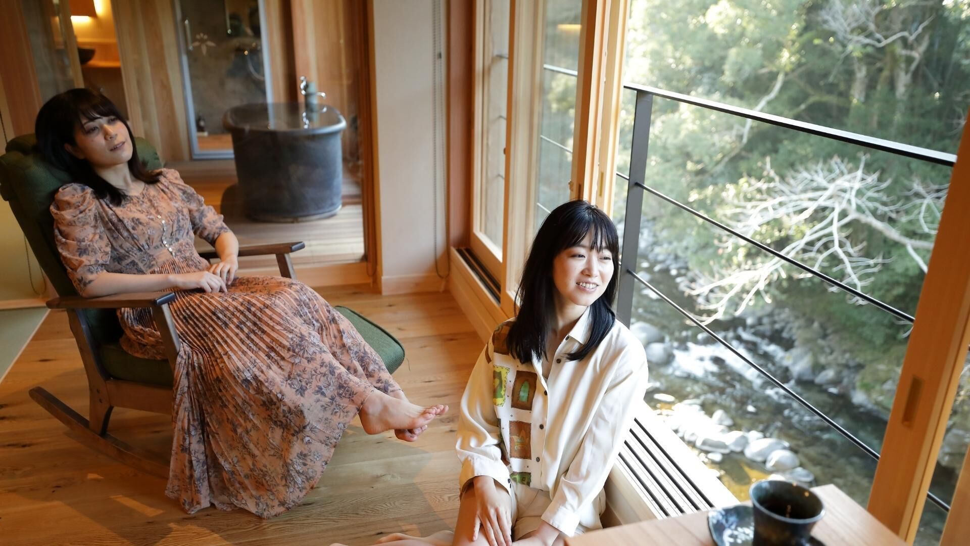 Two people seated in a room with a large window, a wooden floor, and a visible fireplace.