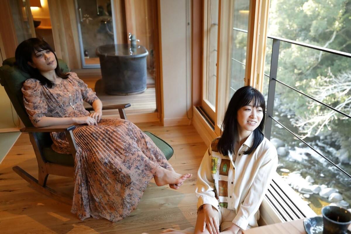 Dos personas sentadas en una habitación con un ventanal grande, piso de madera y una chimenea a la vista.