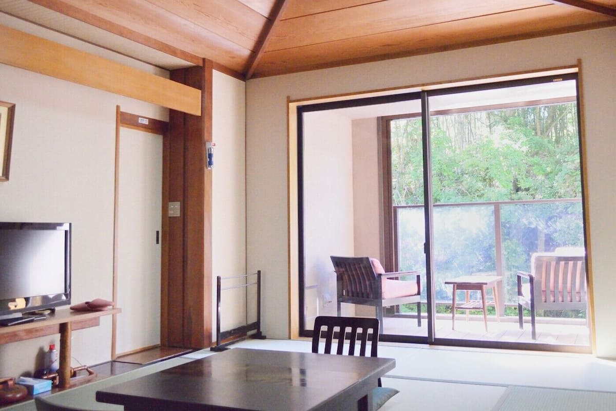 Una habitación con puerta corrediza de vidrio que da a un balcón, una televisión sobre un mueble y una mesa con sillas.
