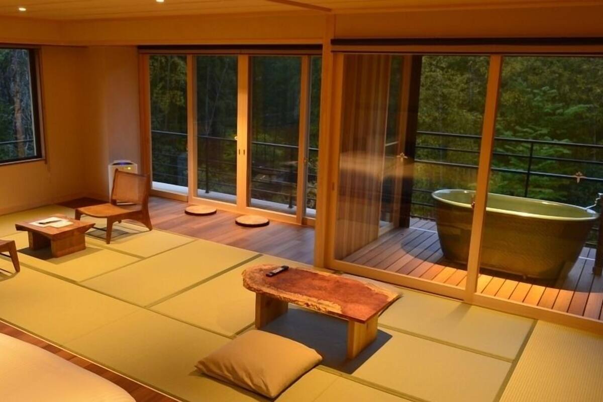 Una habitación con esteras de tatami, una mesa de madera, sillas, una bañera y grandes puertas correderas de vidrio.