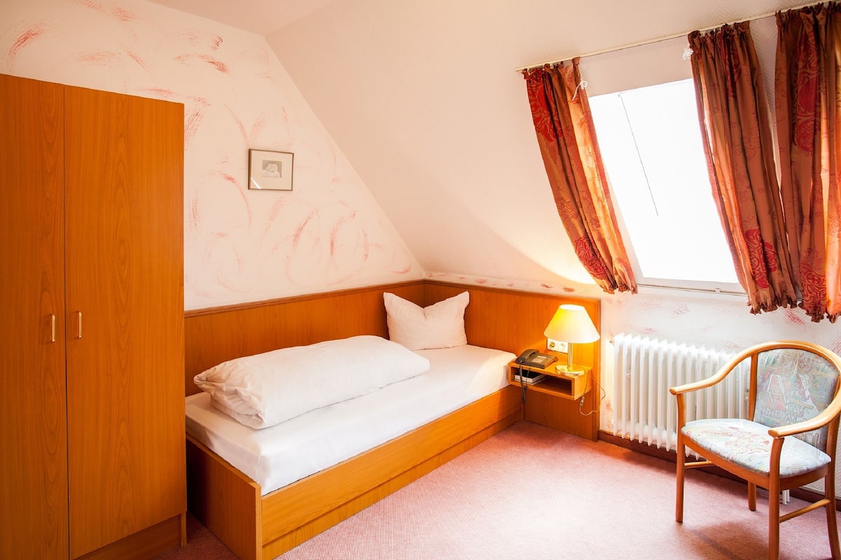 A small, cozy room with a single bed, a wooden wardrobe, a chair, a nightstand with a lamp, and a radiator.
