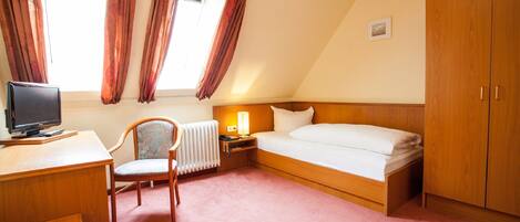 A hotel room with a bed, a desk with a computer, a chair, a radiator, and a wardrobe.