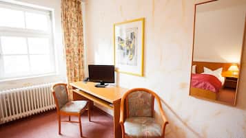 Ein Hotelzimmer mit einem Holztisch, zwei Stühlen, einem Computer, einem Bett mit roter Decke, einem großen Spiegel und einem Fenster mit Vorhängen.