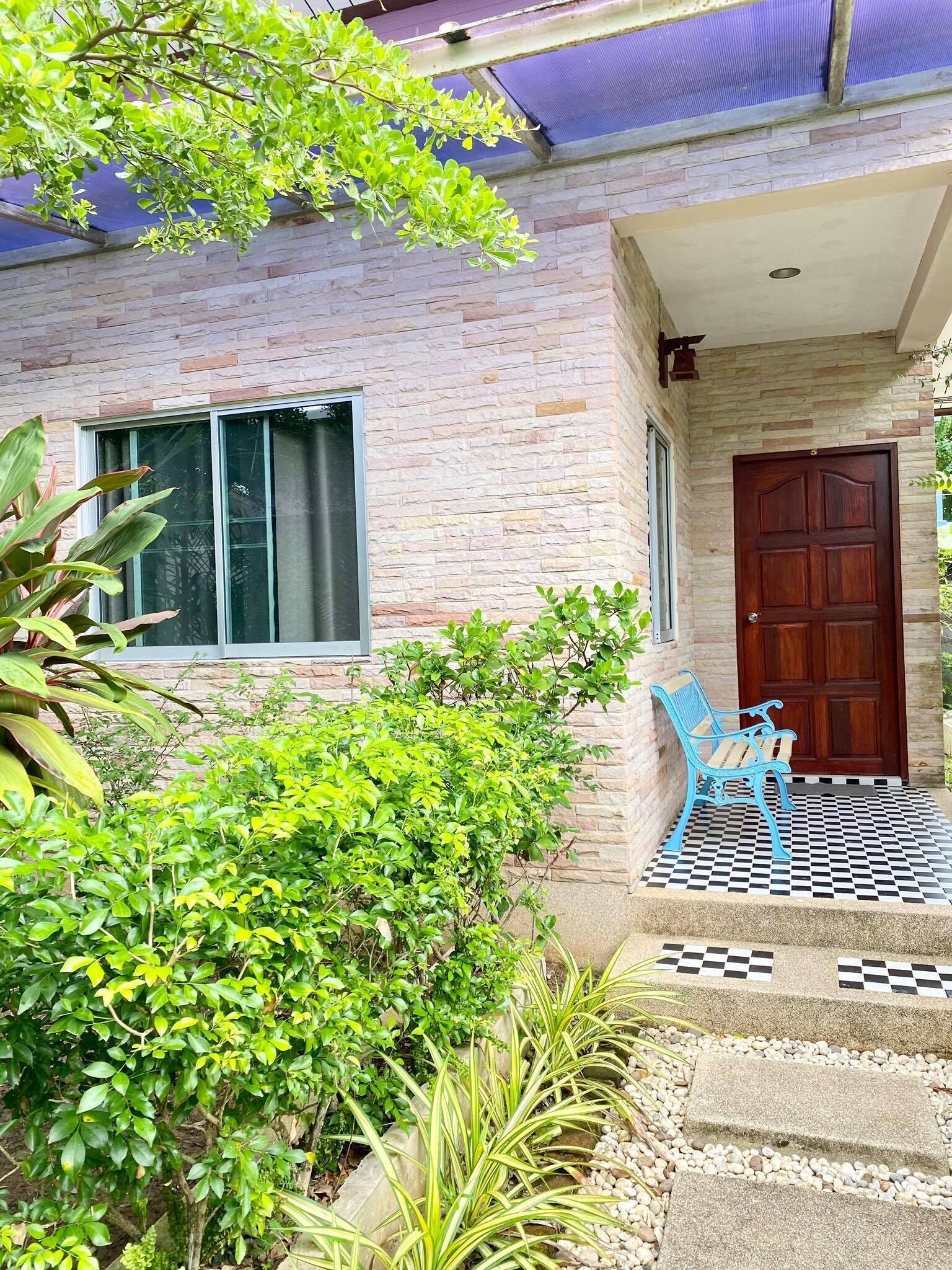 Uma casa com fachada de pedra, uma janela grande, uma porta de madeira e um caminho quadriculado que leva a um banco azul.
