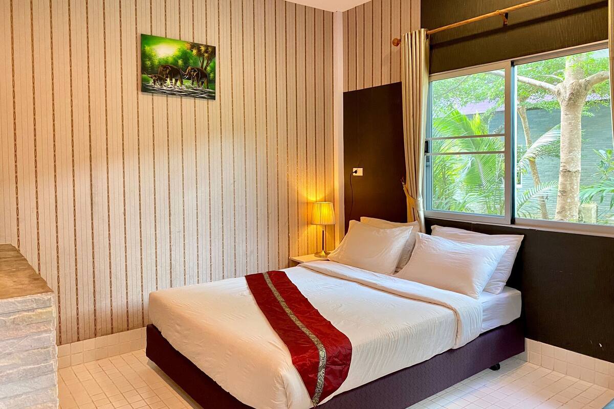 A hotel room with a bed, a nightstand, a lamp, a window with a view of greenery, and a painting on the wall.