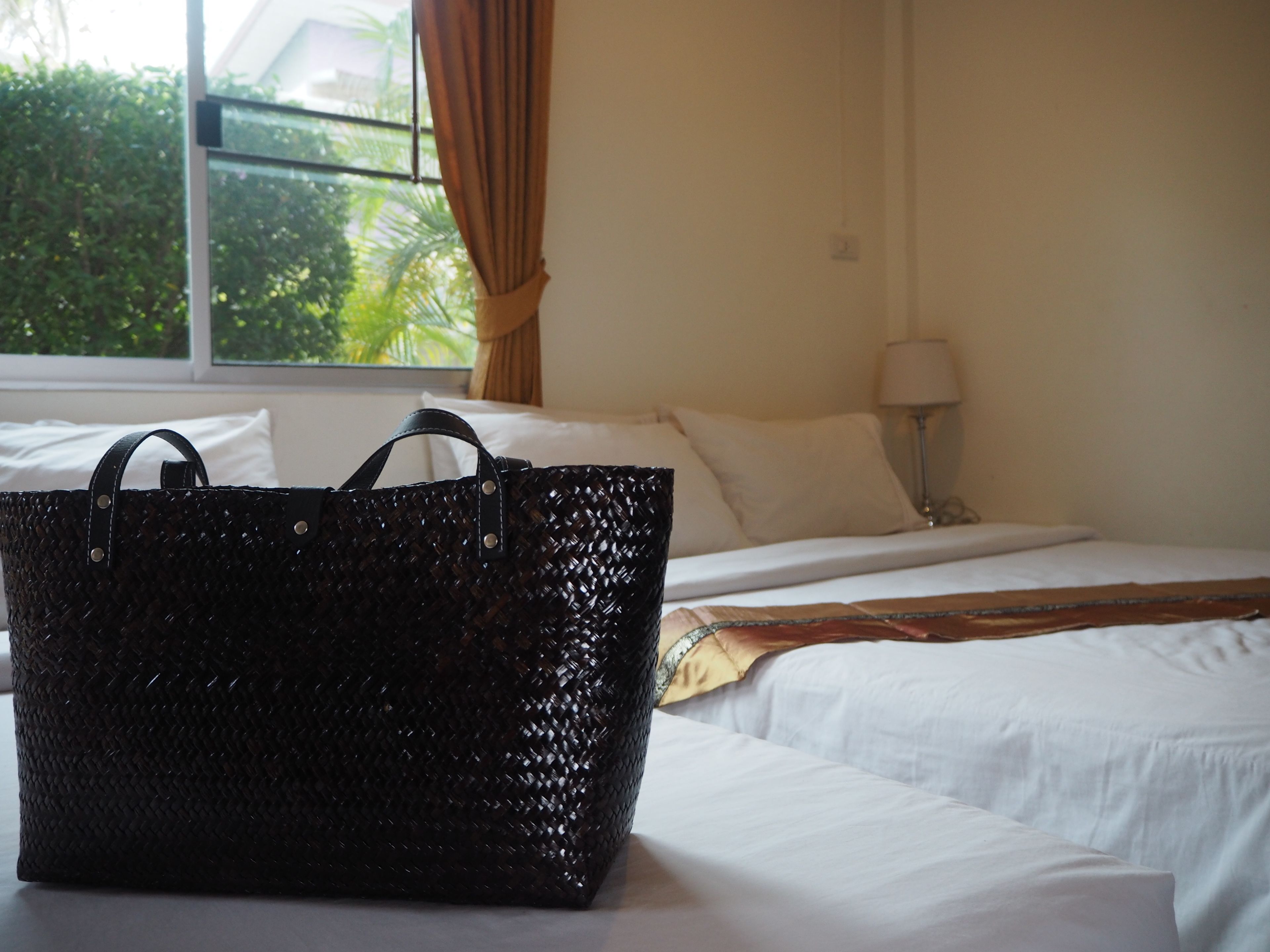 A hotel room with a bed, a window with curtains, and a black woven bag on the bed.